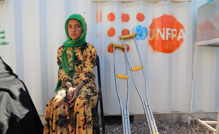 Una niña con vestida de amarillo y con pañuelo verde sentada en una silla fuera de una pared metálica con el logotipo del UNFPA. A su lado hay dos muletas.